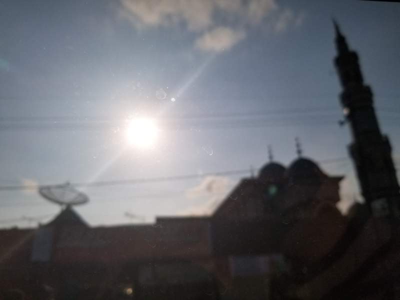 A mosque, slightly out of focus with a large satelite dish to the left & the midday sun shining in the sky above.